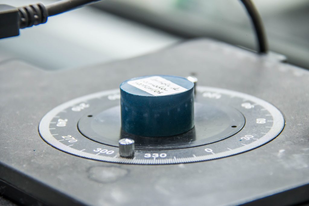 A imagem mostra um equipamento de medição de dureza, possivelmente utilizado para testes como Rockwell (HRC), Brinell ou Vickers. O dispositivo apresenta um disco graduado com marcações angulares e um corpo cilíndrico azul com uma etiqueta identificadora. O cenário sugere um ambiente de laboratório ou controle de qualidade, onde materiais são analisados para determinar sua resistência mecânica.